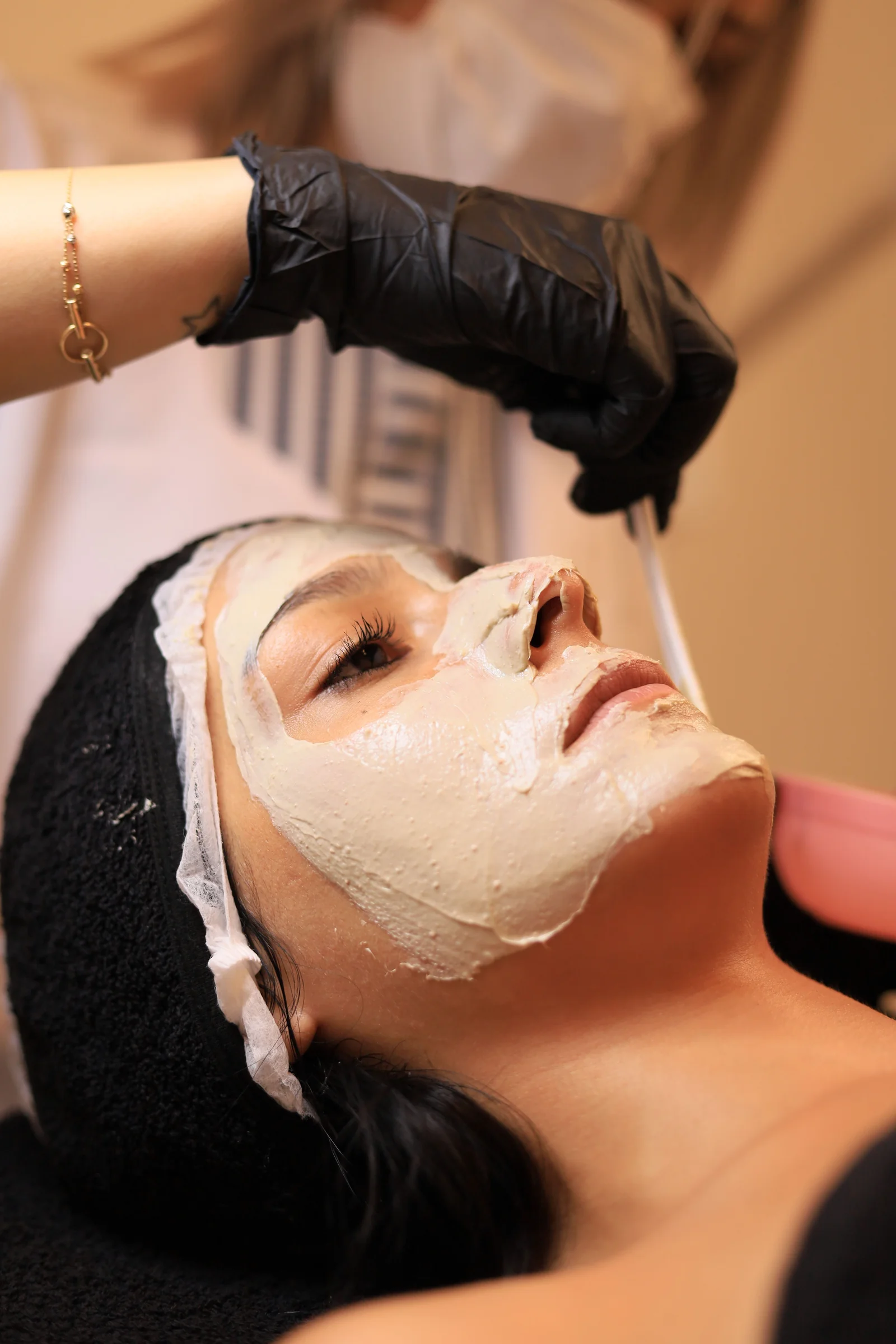 A woman relaxing with eyes closed as gloved hands apply a cream facial mask