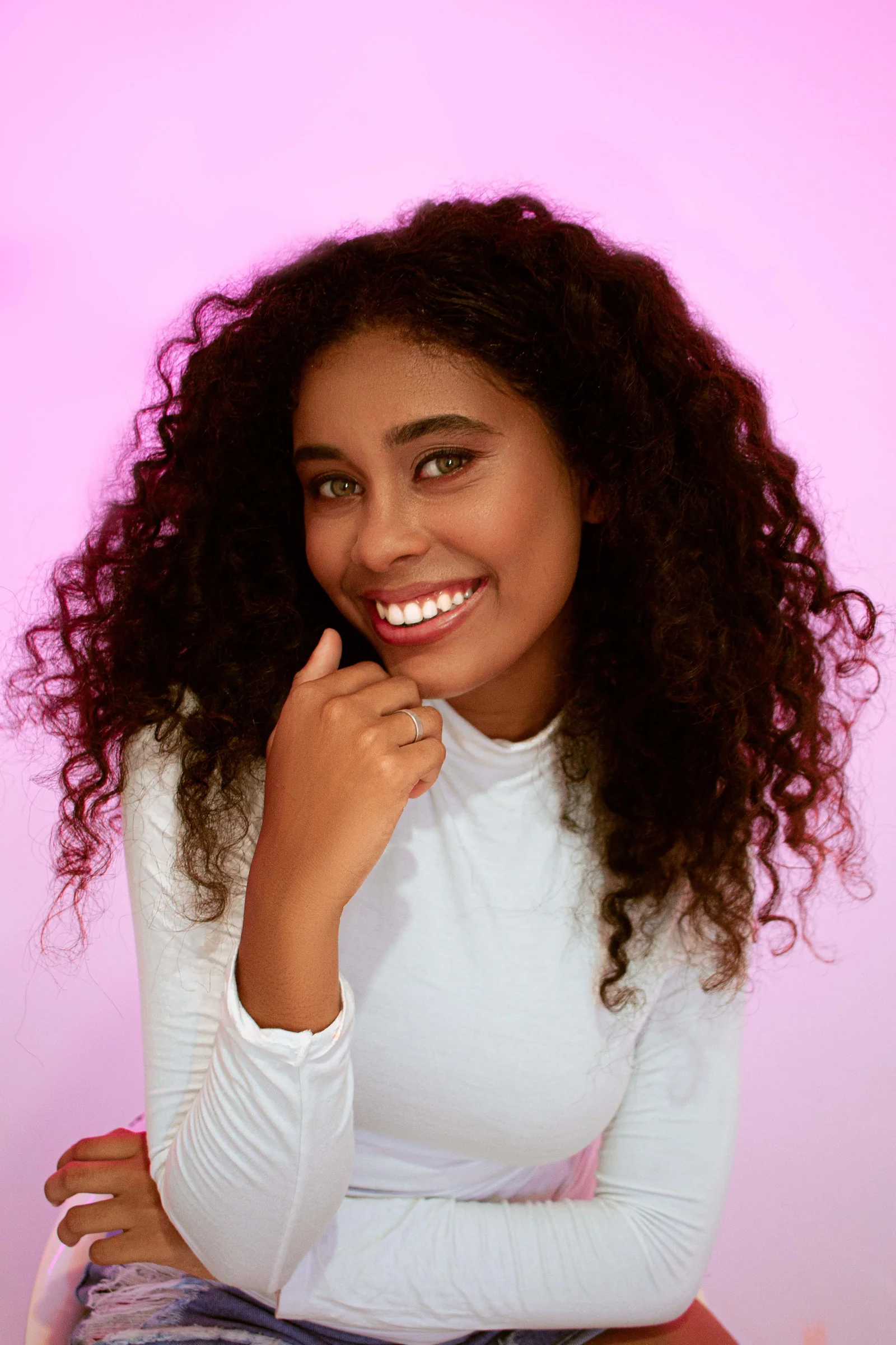 A smiling woman with curly hair in a white top against a pink backdrop