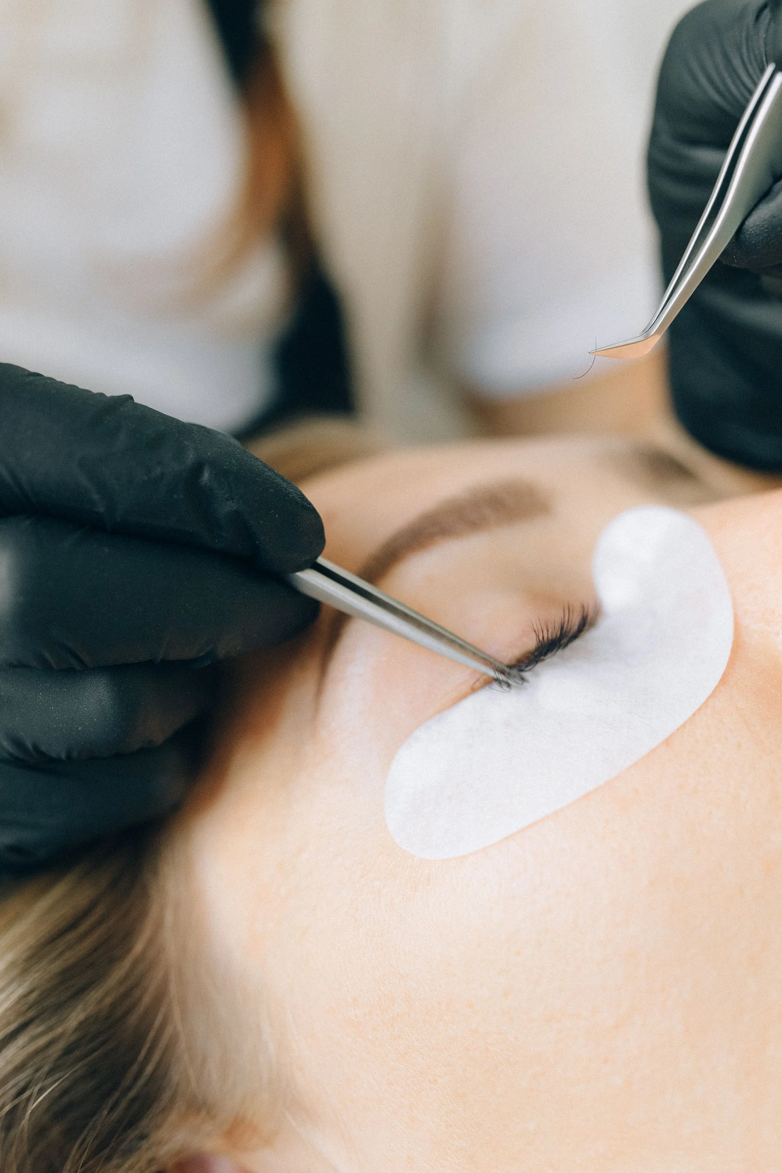 A black-gloved technician applying lash extensions with tweezers